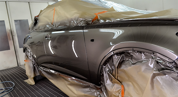 A vehicle in a paint booth with its windows, wheels, and trim masked off using plastic sheeting and tape, showing a freshly painted glossy black surface.