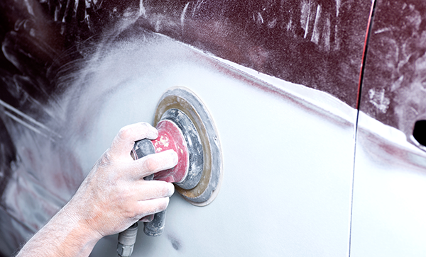 Close-up of a person’s hand using an electric sander to smooth the surface of a car door during auto body repair. The car is partially sanded, with white dust covering the area.